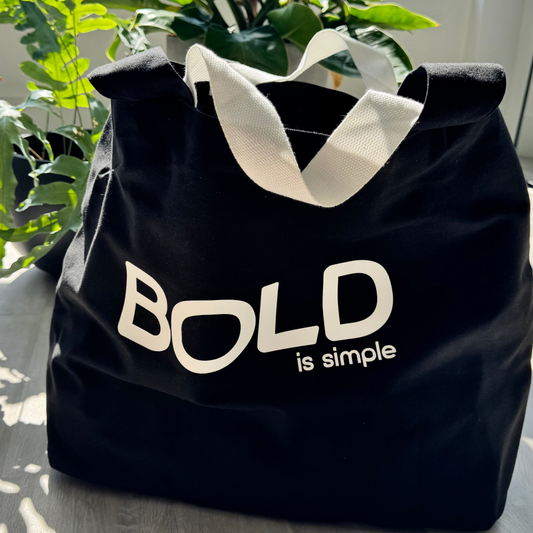 Black tote bag with 'BOLD is simple' text on a wooden surface with plants in the background